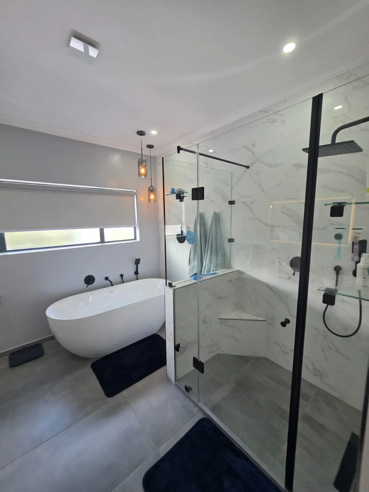 Freestanding bath beside a frameless glass shower enclosure with marble-look wall tiles and black fittings