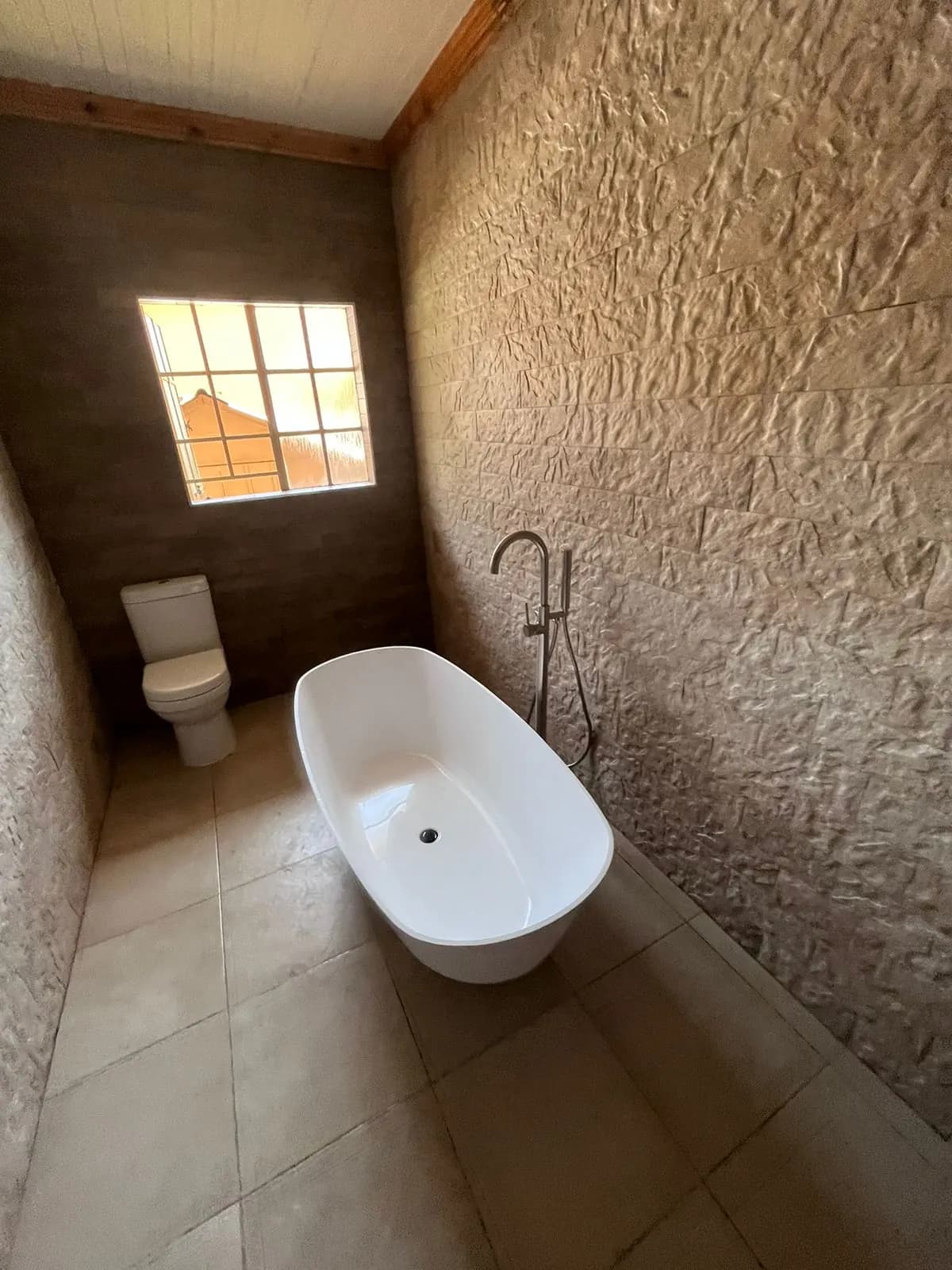 Freestanding bath beneath a window with textured stone-look feature wall