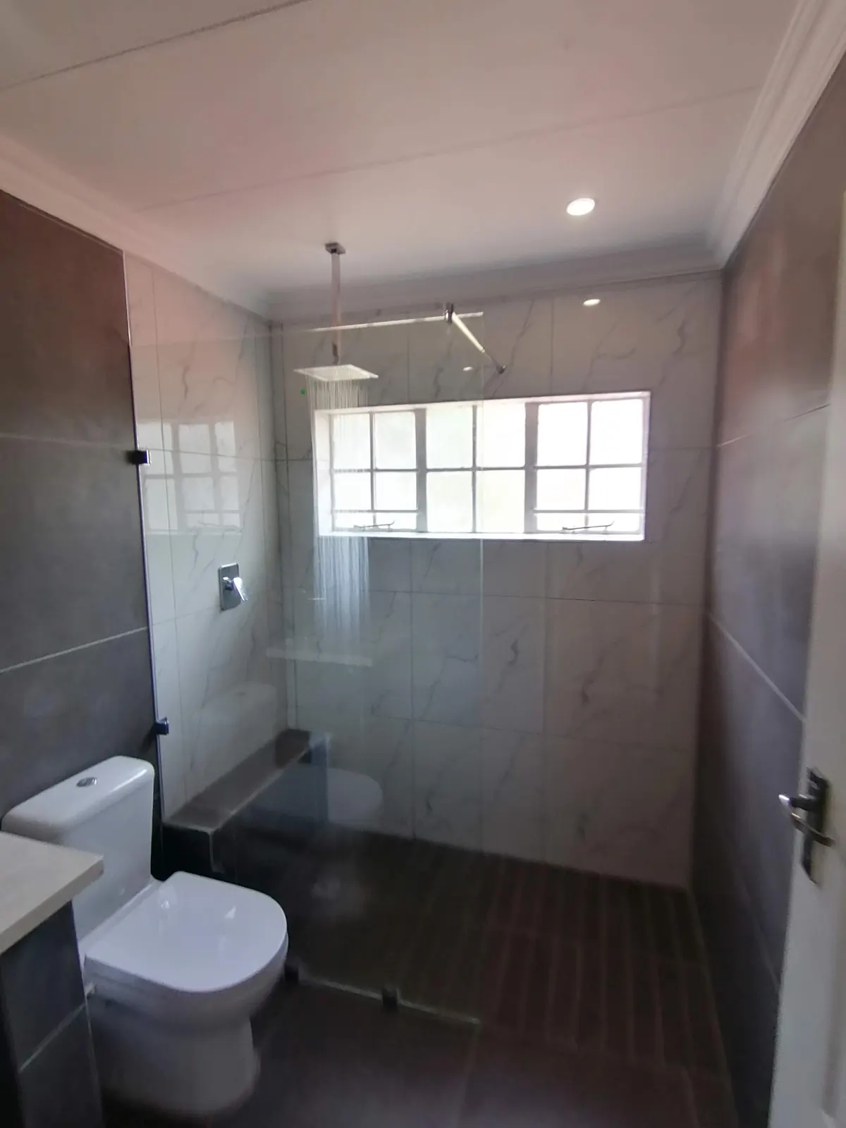 Walk-in shower with ceiling-mounted rain head, frameless glass panel, marble-look wall tiles and tiled bench