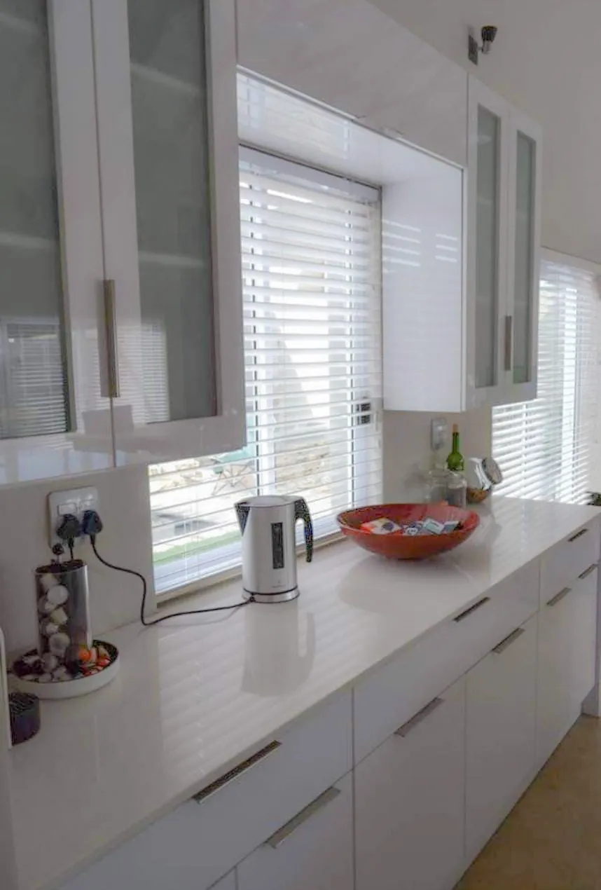 White gloss kitchen with a long stone worktop, handle-less drawers and frosted-glass wall units around the window